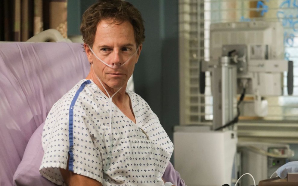 Greg Germann, caracterizado como Tom Koracick, está na cama de um hospital, com tubos saindo de seu nariz e aparelhos médicos ao seu lado em cena de Grey's Anatomy