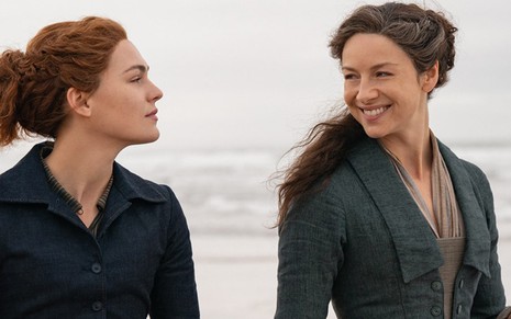 Sophie Skelton (Brianna) olha para Caitriona Balfe (Claire), que sorri, enquanto caminham em uma praia em cena de Outlander
