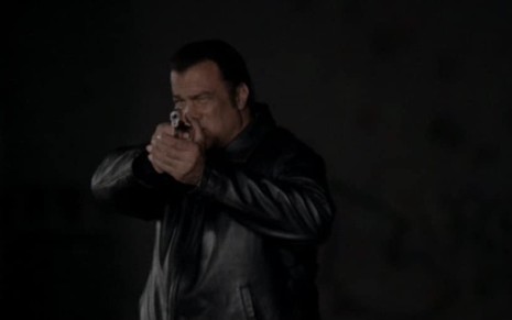 Steven Seagal como o ex-policial Matt Conlin em O Jogador, vetisdo com uma jaqueta de couro preta, ele aponta uma arma.