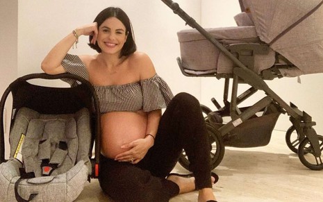 A atriz Sthefany Brito ainda grávida, sorrindo, sentada no chão ao lado de uma cadeirinha de bebê e um carrinho de bebê