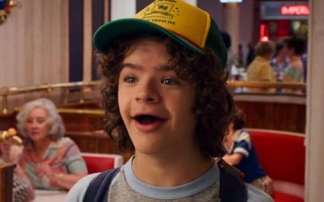 Com a boca aberta de felicidade e usando um boné, Gaten Matarazzo aparece dentro de uma sorveteria em cena de Stranger Things