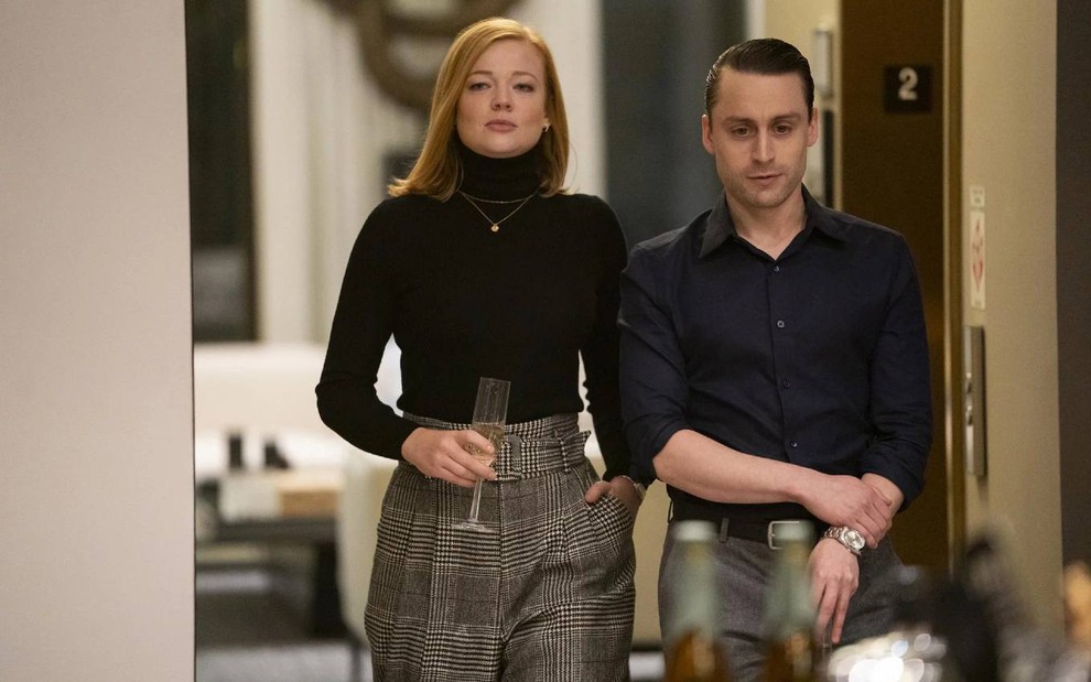 Os atores Sarah Snook e Kieran Culkin lado a lado, com expressões sérias, ela com taça na mão, em cena de Succession