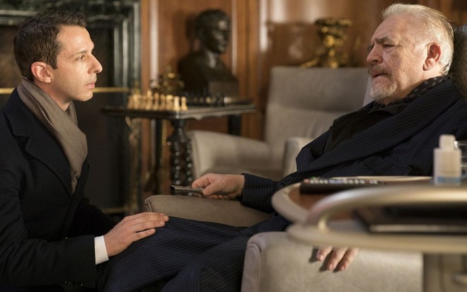 Agachado, Jeremy Strong olha para Brian Cox, sentado em um poltrona, em cena da série Succession