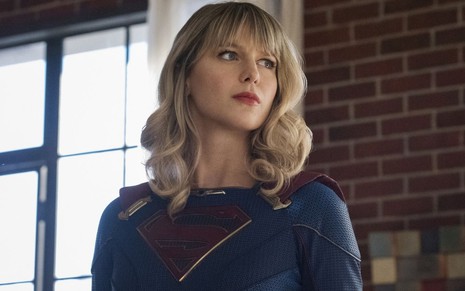 Dentro de casa, com uma parede com tijolos à mostra, Melissa Benoist vestida de Supergirl aparece em imagem da série homônima