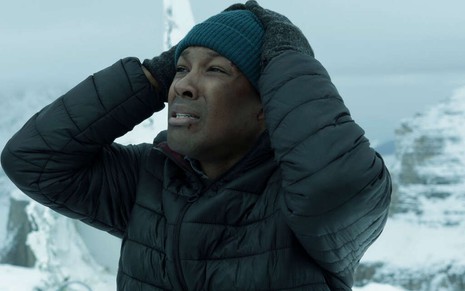 Com casaco de frio e uma touca de frio, de pé em um lugar frio, Corey Hawkins coloca as mãos na cabeça na série Survive