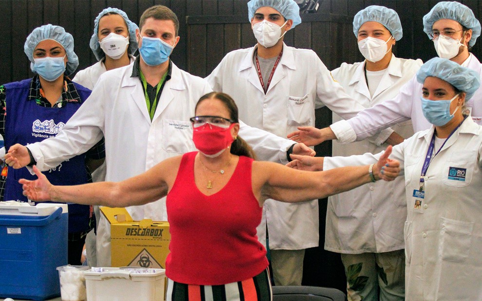Com uma blusa vermelha, que combina com a máscara, Susana Vieira posa de braços abertos na frente de sete profissionais de saúde paramentados com máscaras brancas, jalecos brancos e toucas azuis