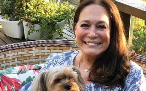 A atriz Susana Vieira no lado externo de sua casa, sorrindo, com um cachorro no colo