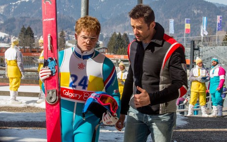 Taron Egerton e Hugh Jackman caminham em uma pista de esqui em cena do filme Voando Alto (2016)