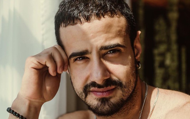 Tarso Brant de barba, corrente no pescoço, sem camisa, com o rosto refletido no sol