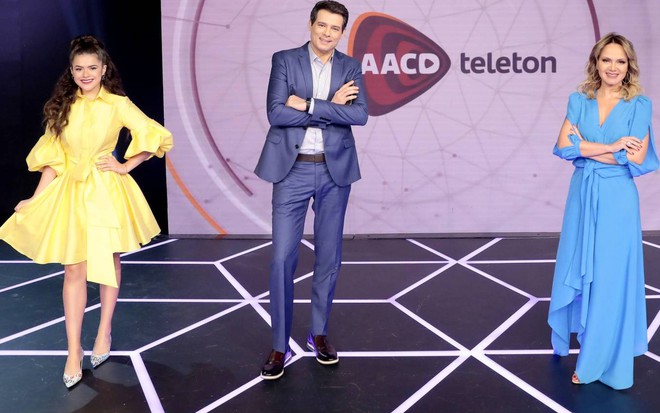 Maisa Silva, Celso Portiolli e Eliana em estúdio do SBT, posam para câmera sorrindo, com logo da AACD num telão atrás