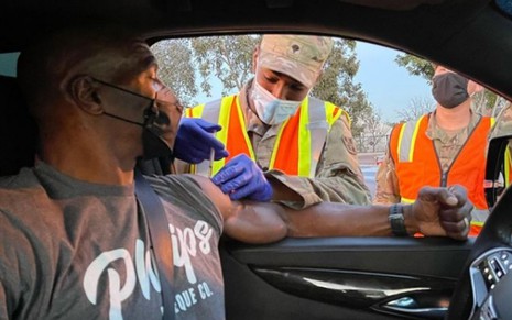 Terry Crews, usando uma máscara preta, recebe vacina contra Covid-19 de militar dentro de seu carro enquanto usa camisa cinza
