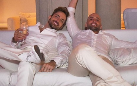 Imagem de Thales Bretas e Paulo Gustavo deitados em sofá com roupas brancas