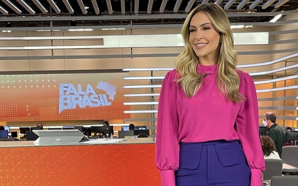 Thalita Oliveira com uma blusa rosa e uma calça roxa, posando de lado, no cenário do telejornal Fala Brasil, da Record