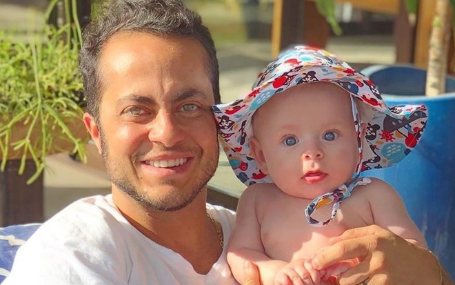 Thammy Miranda de camiseta branca segurando o filho, Bento, no colo. O menino está de sunga azul e chapéu colorido
