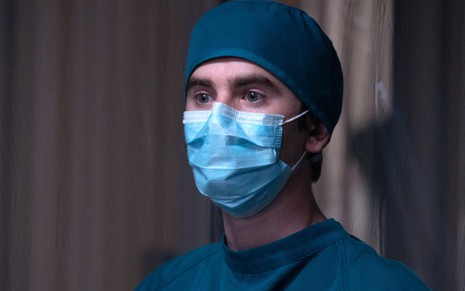 Dentro de um hospital, Freddie Highmore aparece de máscara cirúrgica que cobre nariz e boca em cena de The Good Doctor