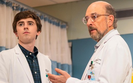 Perto de um leito hospitalar, Freddie Highmore e Richard Schiff conversam vestidos de médicos em The Good Doctor