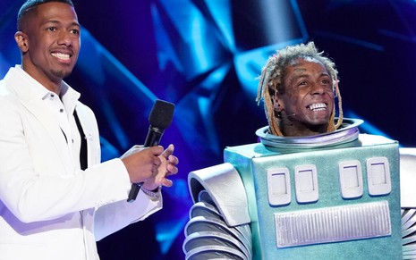 Com uma roupa branca e sorridente, Nick Cannon aparece ao lado do rapper Lil Wayne, fantasiado de robô, no palco de Masked Singer