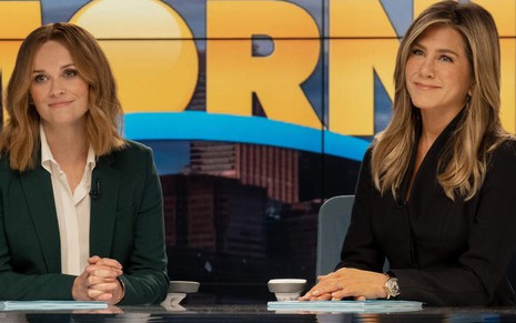 Em uma bancada de telejornal, as atrizes Reese Witherspoon e Jennifer Aniston sorriem timidamente em cena de The Morning Show