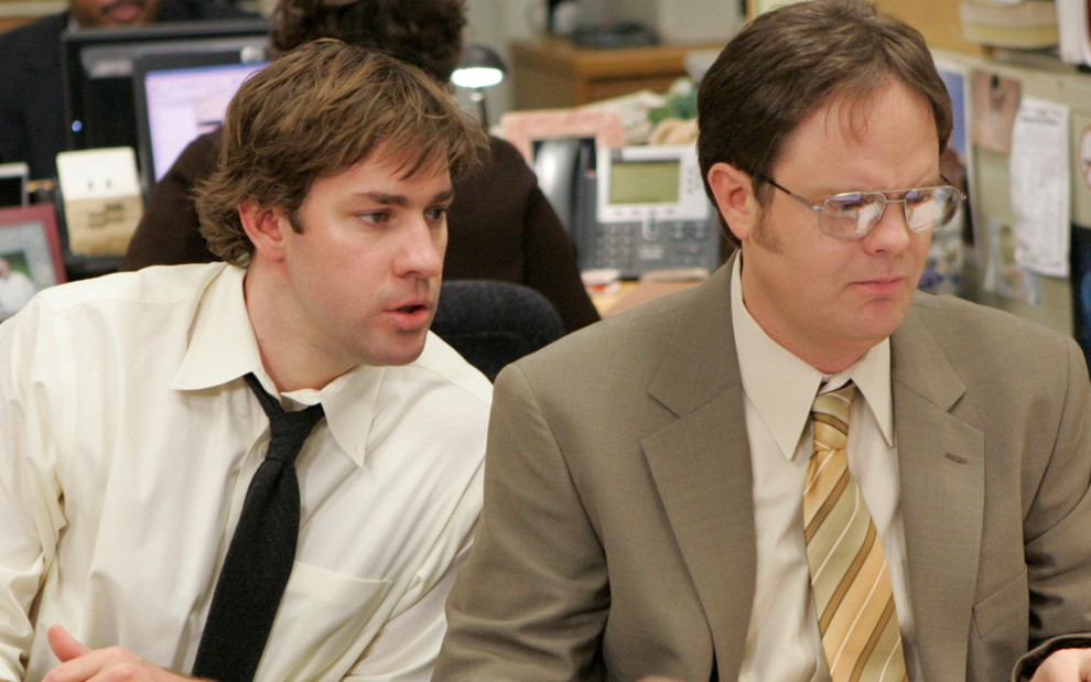 De camisa branca e graveta preta, John Krasinski observa Rainn Wilson mexendo em um computador na série The Office