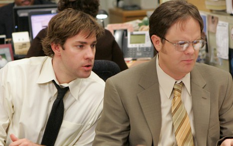 De camisa branca e graveta preta, John Krasinski observa Rainn Wilson mexendo em um computador na série The Office