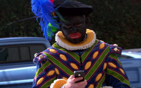Em The Office, Mark Proksch veste uma roupa colorida e enquanto mexe em um celular e aparece com o rosto pintado de preto