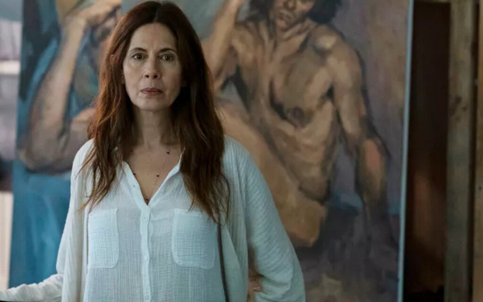 Com um cabelo que ultrapassa os ombros, Jessica Hecht aparece em um ateliê com pinturas de homens nus ao fundo, em The Sinner