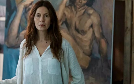 Com um cabelo que ultrapassa os ombros, Jessica Hecht aparece em um ateliê com pinturas de homens nus ao fundo, em The Sinner