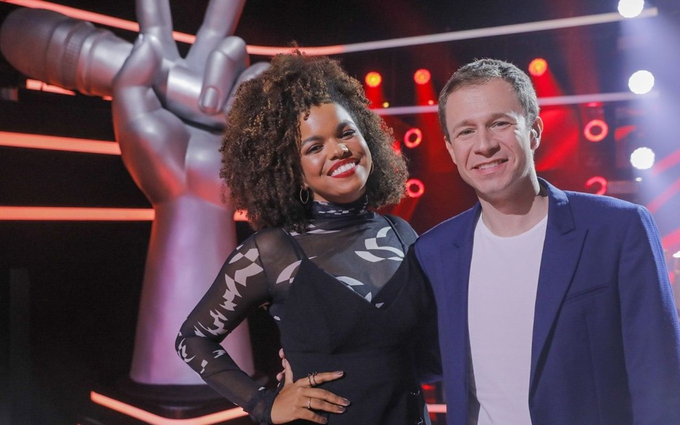 Sorridentes, Jennifer Nascimento e Tiago Leifert posam no cenário do The Voice Brasil