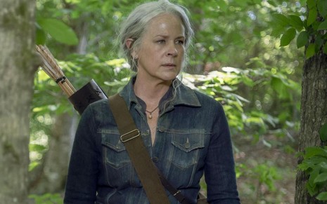 Entre árvores no meio de uma floresta, a atriz Melissa McBride aparece de casaco jeans e com uma arma nas costas em The Walking Dead
