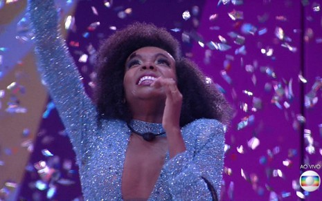 Thelma Assis comemora o prêmio de R$ 1,5 milhão no BBB20; ela ergue os braços com papel picado roxo ao fundo
