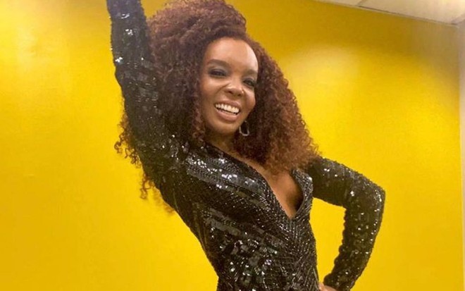 Imagem de Thelma Assis, de cabelo solto e vestido brilhante, com um fundo amarelo
