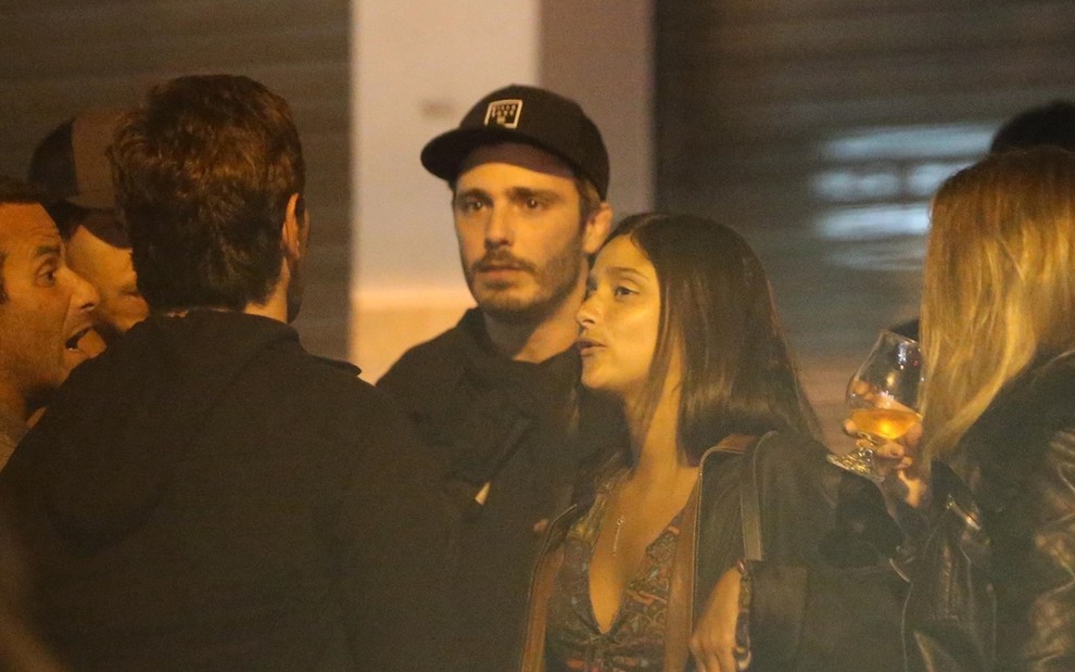 De bonê e roupa de frio, o ator Thiago Rodrigues está rodeado de amigos conversando de pé em um bar do Rio de Janeiro