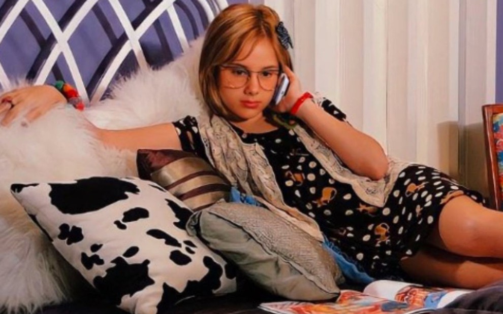 A atriz Clara Tiezzi deitada numa cama, falando ao celular em cena como Mabi de Ti Ti Ti