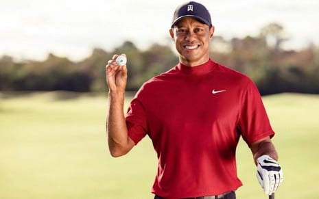 O golfista Tiger Woods, de boné e camisa vermelha, mostra uma bolinha enquanto sorri para a câmera