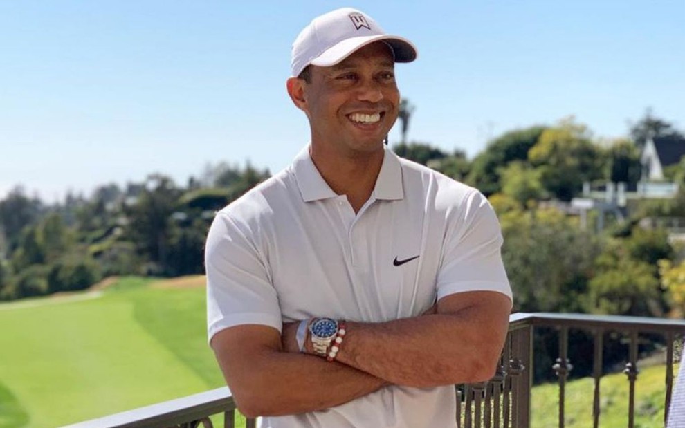 Tiger Woods no campo de golfe, sorrindo, de camiseta e boné brancos e os braços cruzados