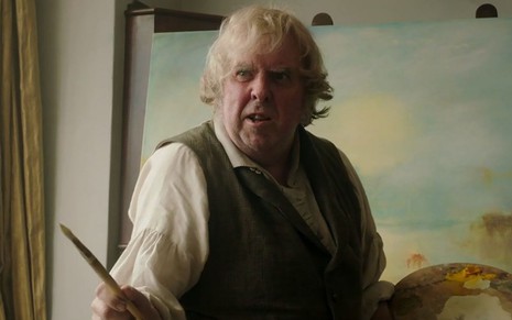 O ator Timothy Spall aparece em frente a uma tela de pintura; ele segura um pincel em cena do filme Sr. Turner (2014)