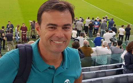 Tino Marcos com a camiseta azul da Globo/SporTV, mochila nas costas, em um estádio de futebol