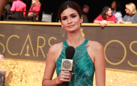 A modelo e apresentadora Carol Ribeiro usa um vestido esverdeado e segura o microfone da TNT no tapete vermelho do Oscar