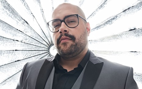 Tiago Abravanel, de paletó cinza com detalhes em preto e usando óculos, em frente a um fundo estilizado