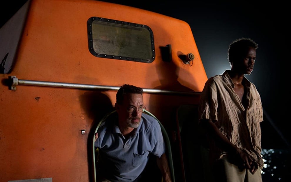 Tom Hanks saindo de uma embarcação marinha laranja e Barkhad Abdi na parte externa da mesma embarcação
