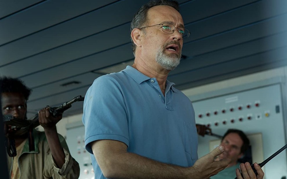 Tom Hanks é sequestrado em cena de Capitão Phillips (2013); ao fundo um ator aparece apontando uma arma para ele
