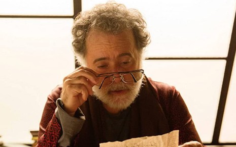 Tony Ramos segurando um óculos na frente dos olhos, enquanto lê um papel