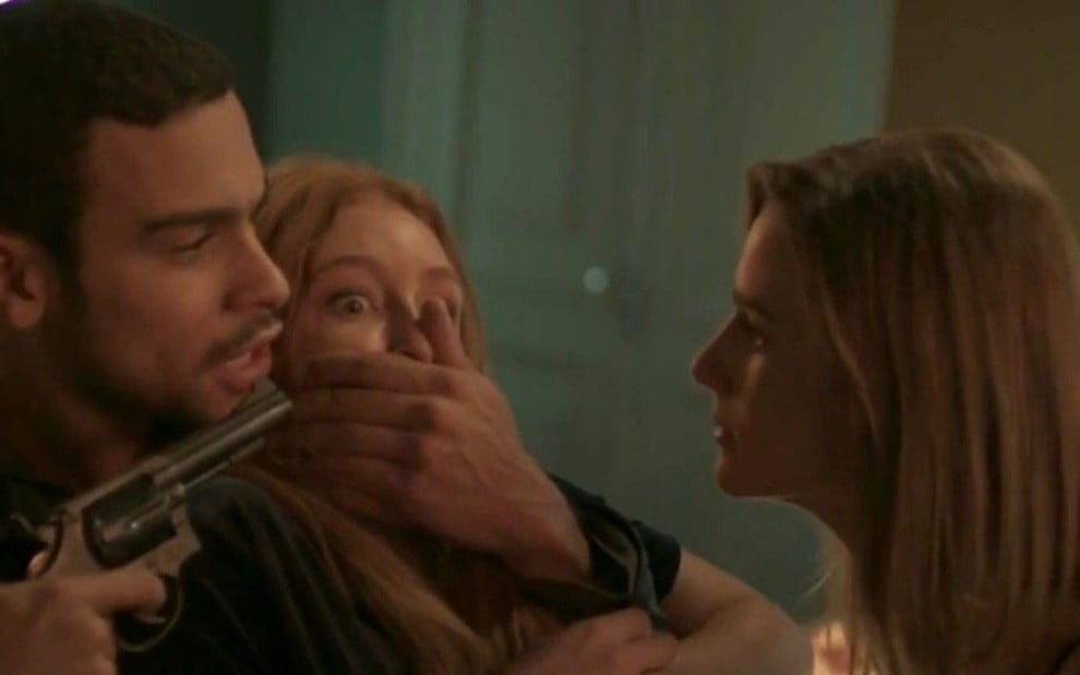 Jacaré (Sergio Malheiros) tapa a boca de Eliza (Marina Ruy Barbosa) e aponta uma arma na cabeça dela; de frente pros dois Sofia (Priscila Steinman), com expressão de raiva