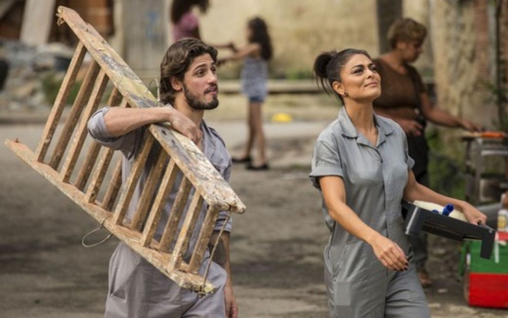 Rafael (Daniel Rocha) e Carolina (Juliana Paes) usam macacões cinza em cena de Totalmente Demais; ele carrega uma escada, e ela um baldinho de tinta