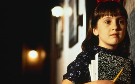 Personagem Matilda, uma garota com franja e tiara de laço, segurando livros e um lápis em um corredor