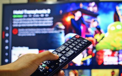 Imagem da mão de uma pessoa segurando um controle remoto com uma smart TV ligada ao fundo