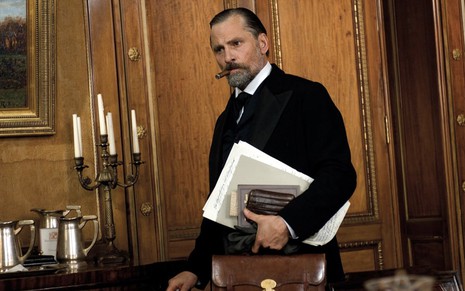 O ator Viggo Mortesen como Freud segura uma pasta com documentos com o braço direito e tem um charuto nos lábios, ele está em um escritório com candelabros ao fundo em cena de Um Método Perigoso