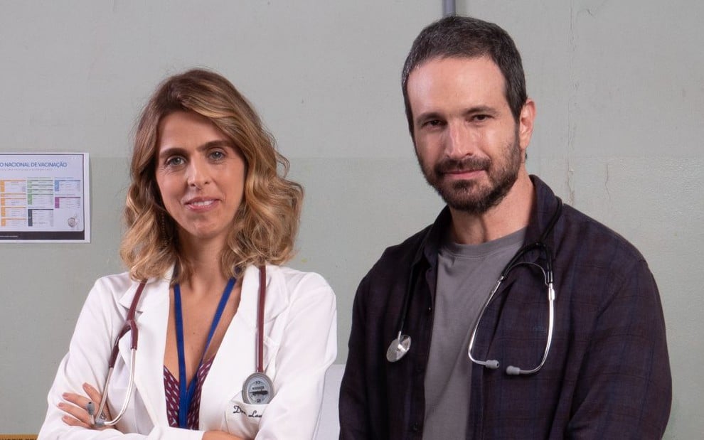 A atriz Ana Petta e o ator Caco Ciocler posam para foto de divulgação, com roupas de médico e estetoscópios no pescoço, no hospital cenográfico de Unidade Básica
