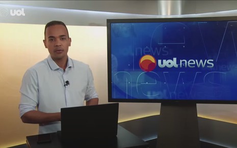O jornalista Diego Sarza no estúdio do UOL ao lado de um monitor em que se vê o logotipo do UOL News