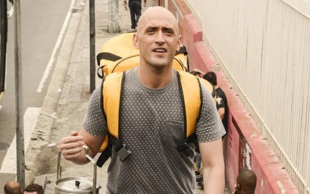 O ator Paulo Gustavo usa uma camiseta cinza e uma mochila amarela em cena como Valdomiro de Vai Que Cola - O Filme